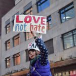 Una bambina con un piumino blu tiene in alto un cartello con su scritto: love not hate