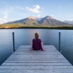 La foto di una persona di spalle, seduta su un pontile di legno che si protende nell'acqua, mentre osservale montagne dall'altra parte del lago, di fronte
