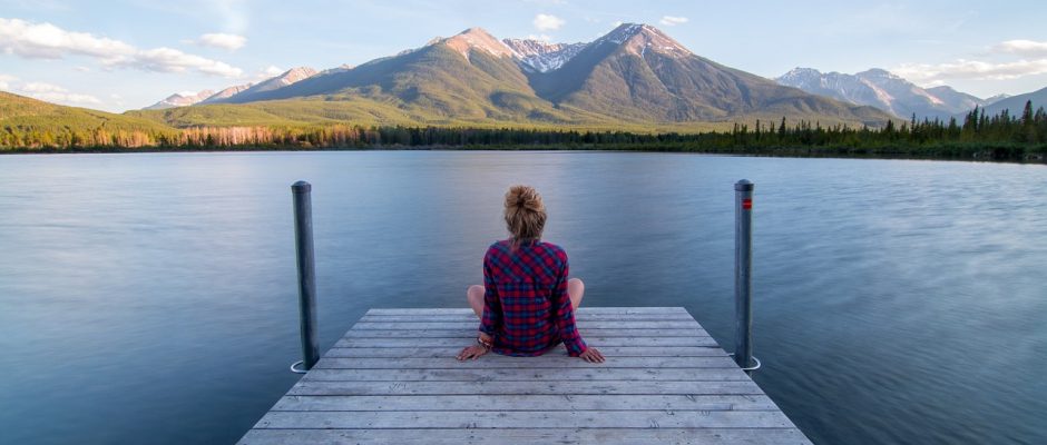 La foto di una persona di spalle, seduta su un pontile di legno che si protende nell'acqua, mentre osservale montagne dall'altra parte del lago, di fronte